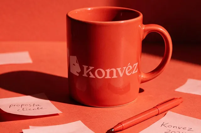 Caneca da Konvéz entre notas de trabalho sobre uma mesa coral.