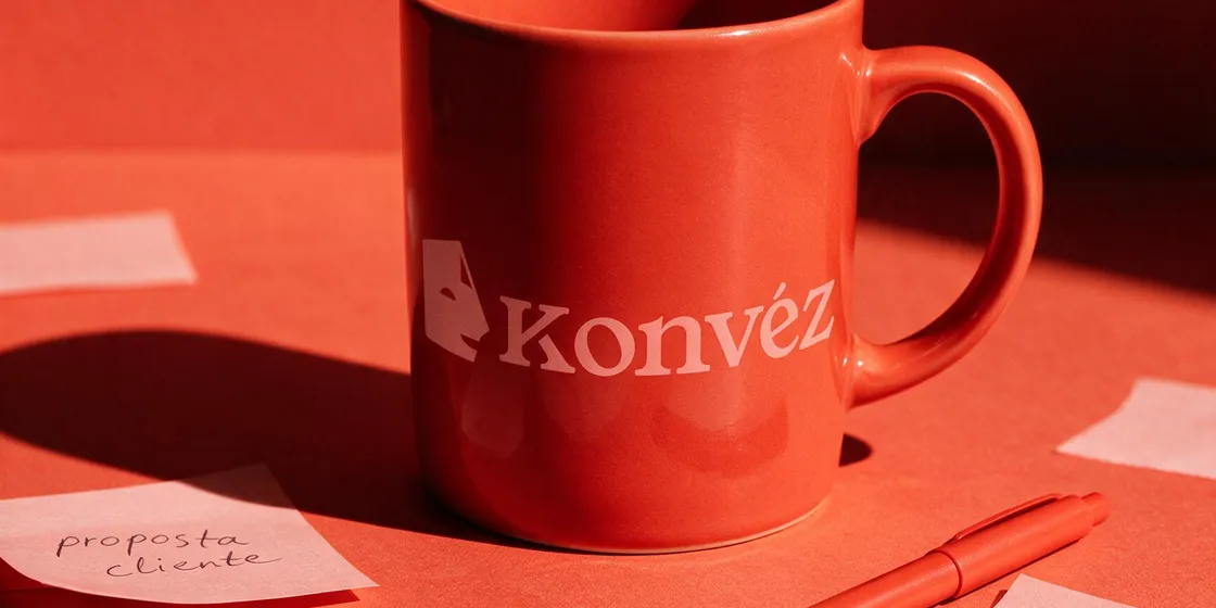 Caneca da Konvéz entre notas de trabalho sobre uma mesa coral.